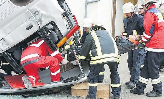 Ein auf dem Dach liegender Kleinwagen, aus dem mit Hilfe von 2 Feuerwehrmännern und 2 Rettungssanitätern eine Person(gespielt) über das Heckfenster
			befreit wird